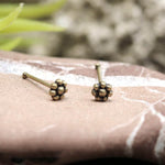 Close-up image of a pair of small, minimalist Kaya Movement FLO Flower Nose Pin Studs in Gold (18 gauge) with floral designs placed on a stone surface. The background has a blurred out-of-focus appearance with hints of green vegetation.