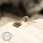 Close-up of a small gold-toned LOTUS Flower Nose Pin Stud in gold, designed by Kaya Movement, placed on a light, textured surface. The minimalist body jewelry shines elegantly. In the background is a blurred logo with the Cyrillic text "КАЧА Модест.