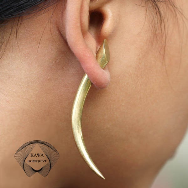 Close-up of a person wearing Kaya Movement's PIKA Fake Talon Gauge Earrings in Gold with a 925 Silver Post. The sleek, elongated design resembles a tusk or horn and loops through the earlobe. The person's ear is in clear focus, with short, dark hair visible in the background, adding a hint of tribal style.