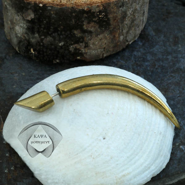 The PIKA Fake Talon Gauge Earrings in Gold with a 925 Silver Post (20 gauge) from Kaya Movement rest gracefully on a white shell. Crafted from yellow brass, these earrings evoke a tribal style with their tapered, curved design ending in a pointed tip. The background is complemented by a textured surface and what appears to be a piece of wood.