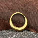 A close-up photo of the AMA Seamless Nose Ring in Gold (9mm) | 18 gauge by Kaya Movement, showcasing its small, circular design with a slight gap. The nose ring is placed against a textured, brownish-red background and is slightly resting on a gray, speckled surface. This minimalist body jewelry piece captures both elegance and simplicity.