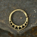 The AYM Seamless Septum Ring in Gold by Kaya Movement is a circular brass body jewelry piece featuring a gold-colored finish. Its semi-circle design showcases small black triangular accents at the bottom, beautifully set against a textured stone background. The seamless ring, available in 18 gauge, includes a small opening at the top for easy insertion and is perfect for various piercing options.