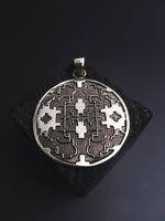 The ICARO - White Brass Handmade Shipibo Icaros Pendant by Kaya Movement features a round, intricately designed pendant with symmetrical, geometric cut-out patterns set against a dark background. The oxidized details emphasize its detailed craftsmanship and artistic design, perfectly capturing the essence of a Shipibo Icaros Pendant.