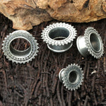 A set of four Kaya Movement TIMO Sacred Lotus Mandala Round Tunnels in Silver, ranging from 3mm to 18mm gauge, are arranged on a textured, dark brown surface. The background includes pieces of natural, rugged brown rock, evoking a tribal plug aesthetic.