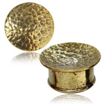 Two gold-tone Hammered Round Saddle Plugs by Kaya Movement, available in 8mm to 16mm gauges. One plug stands on its cylindrical side while the other lies flat, showcasing the front view of their textured surface. These handmade brass earplugs reflect light beautifully, highlighting their shiny and rugged finish.