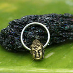 The BUDDHA Silver Ring with Gold Pendant by Kaya Movement is showcased in front of a textured black stone. The ring band, meticulously crafted from gold brass body jewelry, beautifully complements the striking silver tone of the design. The background features a smooth green surface, offering a contrasting backdrop for the ring and stone. Available in 18, 16, or 14 gauge sizes, this piece merges elegance with distinct craftsmanship.