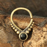 Close-up of a Unique Gold Triangle Septum Ring from Kaya Movement, featuring an 18g size and an 8mm diameter, placed on a wooden surface. The ring boasts an oxidized gold finish with a striking round black onyx stone in the center and elegantly arranged small gold beads symmetrically on either side. The background showcases natural wood textures.