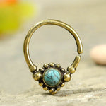 The SYN Seamless Septum Ring in Gold & Turquoise by Kaya Movement is an elegant 18 gauge gold brass body jewelry piece, featuring a small hoop with a decorative closure that showcases a central blue turquoise gemstone surrounded by tiny brass beads. It is displayed on a textured surface with a blurred, colorful background.