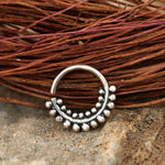 The ELE Seamless Ring in Silver | 18 gauge by Kaya Movement, featuring a semicircular tribal ethnic design with a row of small, round beads along its outer edge, is placed on a textured, earthy surface next to dried, intertwined twigs in the background.