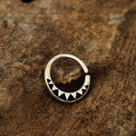 The AYM Seamless Septum Ring in Silver | 18 gauge by Kaya Movement, featuring a series of black triangle shapes and designs along one side, rests on a rough, brown wooden surface.