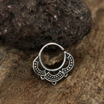 A small Silver Lotus Flower 8mm Septum Ring by Kaya Movement rests on a natural, rough-textured surface. The intricately designed 18g body jewelry features delicate engraved patterns and tiny dot details, adding to its ornate appearance. A dark, rugged stone in the background complements the rustic theme.