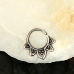 The KAAL Lotus Ring in Silver by Kaya Movement, crafted from delicate 925 Sterling Silver, rests gracefully on a pale, textured surface. This 20-gauge body jewelry piece features an open circular design adorned with five small, teardrop-shaped elements that form a floral pattern. Each teardrop shape is intricately detailed, adding to the ring's elegance.