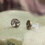 A pair of TREE OF LIFE Labret Studs in Gold by Kaya Movement, crafted from brass and designed with a detailed Tree of Life motif. Displayed on a light beige textured surface, one stud lies flat while the other stands upright, highlighting the intricate detailing beautifully.