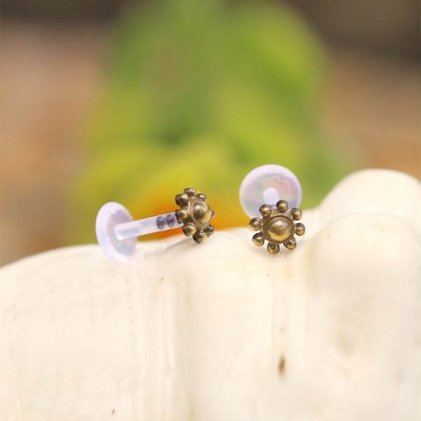 The LUST Flower Labret Stud in Gold, a detailed creation from Kaya Movement, features a central gold bead encircled by intricate petal-like structures. These small, vintage-style earrings are designed with bio-plastic push backs and are beautifully photographed against a softly blurred warm-toned background.