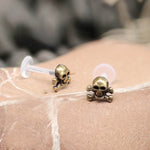Close-up of a pair of PIRA SKULL Labret Stud earrings by Kaya Movement, featuring a punk skull design. The earrings are gold-colored and come with clear plastic backings. They are positioned on a textured surface, possibly stone or marble, with a blurred background.
