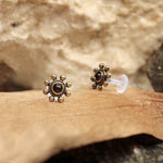 The BEA Flower Labret Stud in Gold & Amethyst from Kaya Movement features a pair of small, intricately designed earrings with a central dark amethyst stone encircled by a sun-like, bronze-colored design that gives off a tribal beaded flower vibe. These stunning 16-gauge earrings are showcased on a wooden surface set against a textured, beige rock background, and come with transparent backings for added elegance.