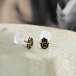 Close-up of a pair of small Kaya Movement earrings, each featuring a minimalist hamsa hand design with a spiral in the palm. The HAMSA HAND Labret Studs, offered in 18 or 16 gauge, are crafted from gold brass and secured with bio-plastic push backs. The earrings rest on a light-colored, textured surface.