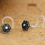 A pair of decorative nose pins with clear hooks is displayed on a wooden surface. The Flower Nose Stud by Kaya Movement features intricately designed floral patterns with a central pearl-like bead and black ornate detailing, showcasing the elegance of Silver 925 craftsmanship.