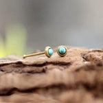 Close-up of two small, round Kaya Movement EDEN nose pin studs featuring turquoise stones set in gold-tone metal. The studs are placed on a textured wooden surface, with a blurred background. These minimalist EDEN nose studs in gold with smooth, polished turquoise stones offer a simple yet elegant look.