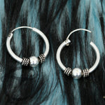 Two SPIA Simple Tribal Hoop Earrings in silver from Kaya Movement, crafted from sterling silver 925 with intricate bead accents, are set against a textured black and blue background. The earrings feature a smooth round bead and twisted wire bead, enhancing their stylish design.