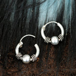 A pair of Kaya Movement's SPIA Simple Tribal Hoop Earrings in Silver (18 gauge) lie on a textured, dark background. Each sterling silver 925 earring features a central silver bead flanked by intricate, coiled metal designs. The slightly open hoops showcase their latch closures perfectly.