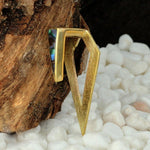 A close-up of Kaya Movement's DIA Minimalist Triangle Ear Hangers in Gold & Abalone Shell, showcasing their geometric design with a triangular shape that loops back on itself. These 0 gauge ear hangers, echoing the allure of body jewelry, are adorned with small iridescent abalone shell inlays. They rest against a textured wood background and are surrounded by white pebbles.