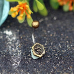 Introducing the Om Chakra Belly Piercing Ring in Gold by Kaya Movement. This 14 gauge belly button piercing showcases a vintage brass design, featuring an "Om" symbol intricately engraved on a circular disc at the bottom, with a round brass ball adorning the top. Expertly crafted with surgical steel for enhanced durability, it is elegantly packaged in beautiful gift packaging. The background includes softly blurred green leaves and delicate orange flowers.