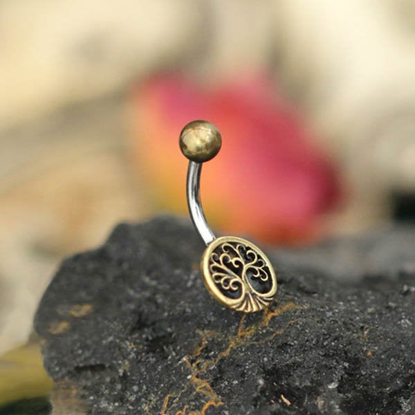 The Tree of Life Belly Piercing Ring in Gold by Kaya Movement is a beautifully curved navel jewelry piece featuring a golden top ball and an intricately detailed tree of life charm at the base. The charm rests gracefully on a dark, textured surface, with a blurred background showcasing earthy tones and hints of red-orange—perfect for elegant gift packaging. This exquisite piece is designed in 14 gauge for timeless sophistication.