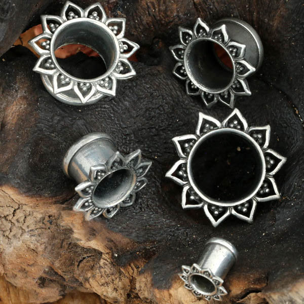 Several Kaya Movement's NIN Sacred Lotus Flower Round Tunnels in silver are displayed on a dark, textured wooden background. The gauges range from 3mm to 18mm and feature an ornate design with detailed petals encircling each tunnel, reminiscent of a Mandala Lotus flower.