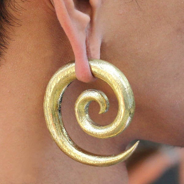 A close-up of a person’s ear adorned with the SPIRAL Minimalist Ear Weights in Gold by Kaya Movement. The gold jewelry starts from the earlobe and loops outward in a smooth, shiny spiral, ending in a pointed tip. The texture of the earring is slightly brushed, giving it a sophisticated look.