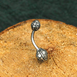 A close-up shot of the Kaya Movement's GEO Globe Belly Piercing Ring in Gold or Silver, featuring two ornate, spherical ends with intricate designs. The 14 gauge navel jewelry is placed on a textured wooden surface against a dark background.
