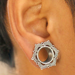 Close-up of a person wearing a Kaya Movement LOTUS Sacred Lotus Mandala Round Tunnel in Silver. The jewelry, situated in an ear lobe, features intricate petal-like details around the circular tunnel in the center. The medium brown skin tone of the ear highlights the beauty of this handcrafted tribal plug, available from 4mm to 25mm gauge.