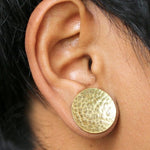 Close-up of a person's ear adorned with a large Hammered Round Saddle Plug in Gold by Kaya Movement. The plug, available in gauges from 8mm to 16mm, covers the earlobe entirely and features a textured, hammered brass surface. The person has short, dark hair.
