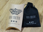 A brown paper pillow box and a black cloth drawstring bag, each labeled with "Kaya Movement," display their email, Facebook, Instagram, and website information. Both items are placed on a wooden surface alongside stunning ethnic body jewelry and an elegant pair of Huggie Hoop Earrings in Silver 925 from Kaya Movement.