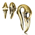 Displayed are three RAVEN SKULL Gothic Ear Weights in 5/8 gauge by Kaya Movement, each sculpted to resemble a metallic bird skull in varying sizes. The intricate detailing includes elongated beaks and hollowed eye areas, capturing realistic avian anatomy with a polished brass finish. They are artfully positioned to showcase different angles and perspectives.