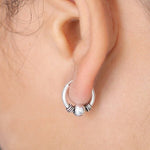 Close-up of a person's ear adorned with a small, circular "Huggie Hoop Earring" from Kaya Movement, featuring decorative ridges and a central bead. The background is blurred, focusing attention on the tribal earring and ear, with some strands of hair visible near the ear.