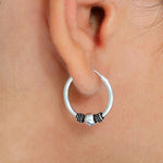A close-up shot features a person's ear adorned with SPIA Simple Tribal Hoop Earrings in Silver by Kaya Movement. These minimalist 18 gauge sterling silver 925 earrings have a smooth, round silver bead at the center, accented by small, coiled black details on either side. The ear and earring appear sharply in focus.