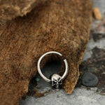 A small SKULL Gothic Septum Ring in Silver by Kaya Movement, crafted in 925 sterling silver with a skull detail, rests on a piece of rough, textured bark against a concrete surface. The intricately designed skull enhances the gothic aesthetic of this 18-gauge ring, making it an edgy and versatile piercing accessory.