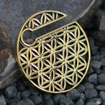 The FLOWER OF LIFE Disc Ear Weights in Gold by Kaya Movement, crafted for 00 gauge ears and featuring a circular cutout adorned with an intricate Flower of Life design, reminiscent of exquisite body jewelry, are placed on a textured, dark rocky surface.