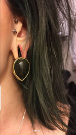 Close-up of a person’s ear adorned with OBI Minimalist Oval Ear Hangers in Gold & Golden Obsidian by Kaya Movement. The person's dark hair partially obscures their ear, and a blurred background is visible.