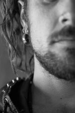 Black and white close-up of a man's face showing only half. He has facial hair, curly hair partially covering an ear adorned with Kaya Movement Silver Futuristic Hoop Earrings Cyberpunk Jewelry. He's wearing a spiked leather garment, and the image focuses on the texture and detail of his beard and accessories, embodying a cyberpunk design.