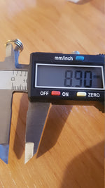 A digital caliper is measuring the width of a small, multi-banded TRIA Septum Ring in Gold by Kaya Movement, which reads 8.90 mm on its electronic display. The measurement is being taken on a wooden surface.