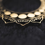 Close-up image of a golden decorative ornament featuring intricate heart-shaped designs. The detailed metalwork, reminiscent of the Lotus Flower Pair Earrings from Kaya Movement, displays curved lines and dots against a dark, textured background. The focus highlights the artistry and fine craftsmanship of this handmade mandala brass jewelry piece.