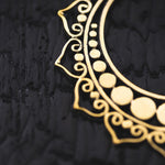 A close-up photograph showcasing the intricate design of the Lotus flower Pair Earrings by Kaya Movement, highlighting the handmade mandala brass pattern against a textured, black background. The gold earrings feature a curved shape adorned with circular and spiral motifs, creating an elegant and ornate appearance.