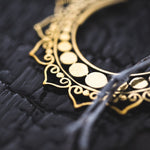 Close-up of an intricately designed gold necklace placed on a dark, textured surface. The necklace features ornamental patterns with circular and swirling elements and a string that appears to be partially draped over it, evoking the elegance similar to Kaya Movement's Lotus Flower Pair Earrings Handmade Mandala Brass Jewelry.