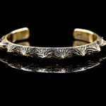 The KRAKO Reptilian Spine Cuff Bangle by Kaya Movement features a rugged, nature-inspired design crafted using the lost wax process. This unisex open cuff bangle, available in gold or black, showcases jagged edges reminiscent of mountain peaks or tree bark. It has an adjustable size and is displayed on a shiny black surface that reflects its intricate details.