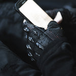 A person wearing DZALA Black Genuine Leather Fingerless Gloves from Kaya Movement, featuring studded knuckles and red accents, holds a smartphone, focusing on the screen. The gloves have visible textured patterns and protective elements on the knuckles. The background is dark and blurred, emphasizing the hands and phone as the central subjects.