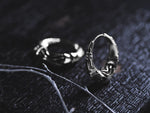 Two intricately designed silver futuristic hoop earrings from Kaya Movement, featuring a cyberpunk aesthetic, placed on a dark textured surface. One earring stands upright, highlighting its detailed side, while the other lies flat and partially blurred in the background. Thin twigs are also visible.