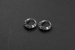 A pair of Kaya Movement's Silver Futuristic Hoop Earrings, designed for the cyberpunk enthusiast and crafted from sterling silver with intricate details, lies on a dark gray surface. Featuring a secure snap latch closure, these unisex earrings are showcased parallel to each other, highlighting their SciFi-inspired craftsmanship.