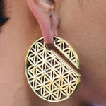 A close-up of a person's ear adorned with Kaya Movement's FLOWER OF LIFE Disc Ear Weights in Gold, designed for 00 gauge. The large, circular gold earring features an intricate geometric pattern that resembles the flower of life and includes a unique design with a small open section. The person's warm-toned skin highlights the earring's exquisite details.